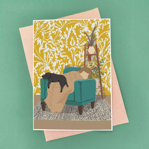 Black labrador sleeping on their dog mum in yellow armchair, with patterned wallpaper background